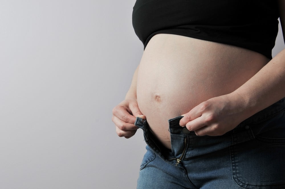 Woman attempting to put on jeans with pregnant belly in the way