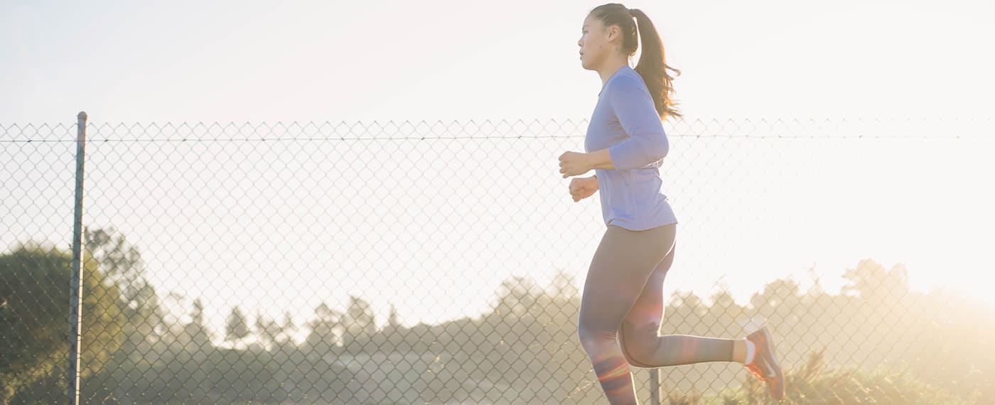 Woman in fitness gear running alongside chain-link fence with the sun in the background