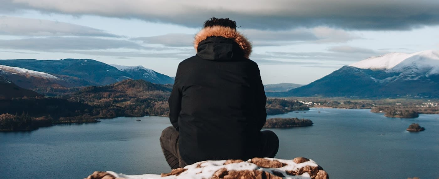 Man sitting on snowy hilltop looking out at lake and snowy mountain tops