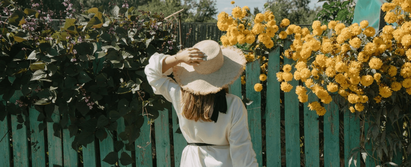 woman with straw hat looking at yellow flowers growing through a wooden fence
