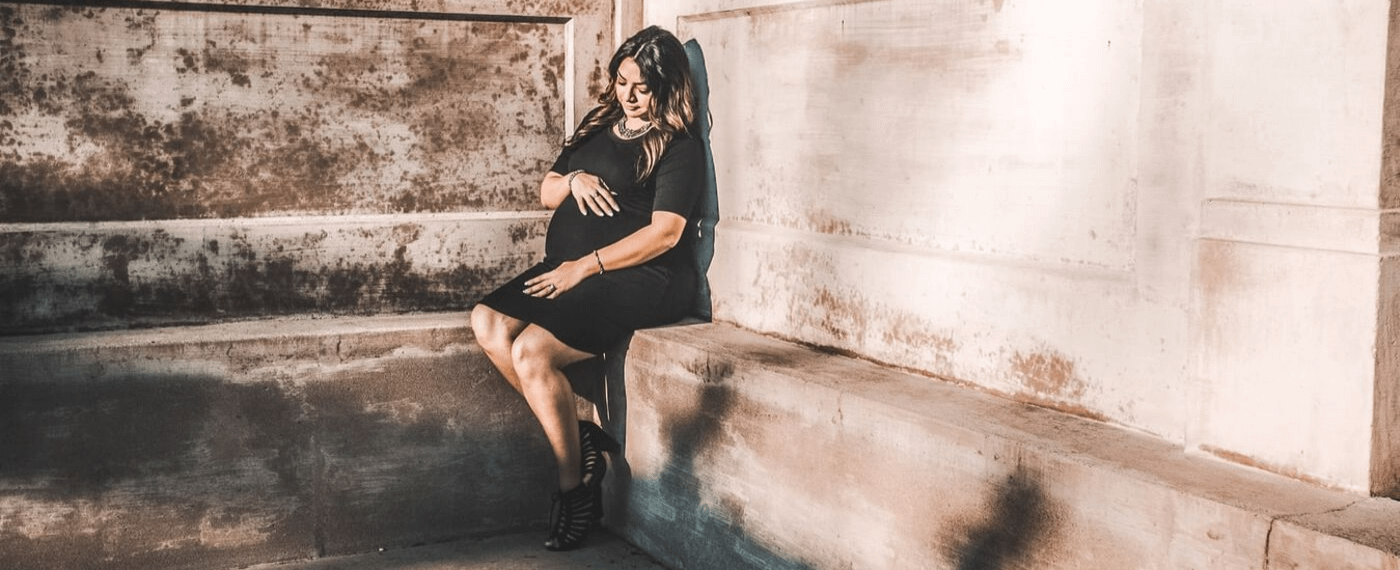 Pregnant woman in black dress sitting on a cement ledge holding her stomach