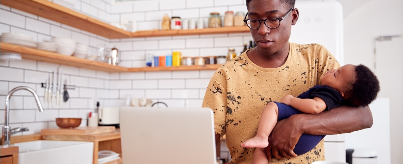 Young father holding newborn son in one arm while working on a laptop