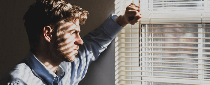 Man looking through blinds of a window with sunlight pouring thorugh