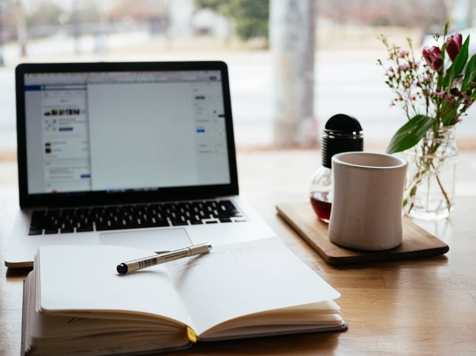 A open notebook and pen sitting in front of a laptop and cup of coffee