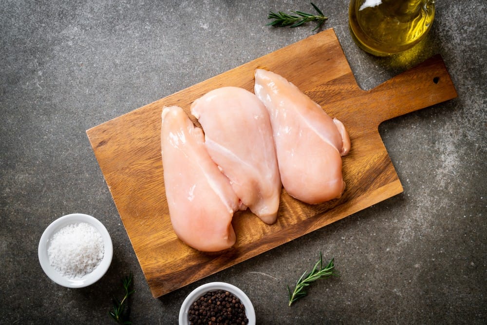 Raw chicken breasts in top of a wood plank with salt, pepper, and oil on the side