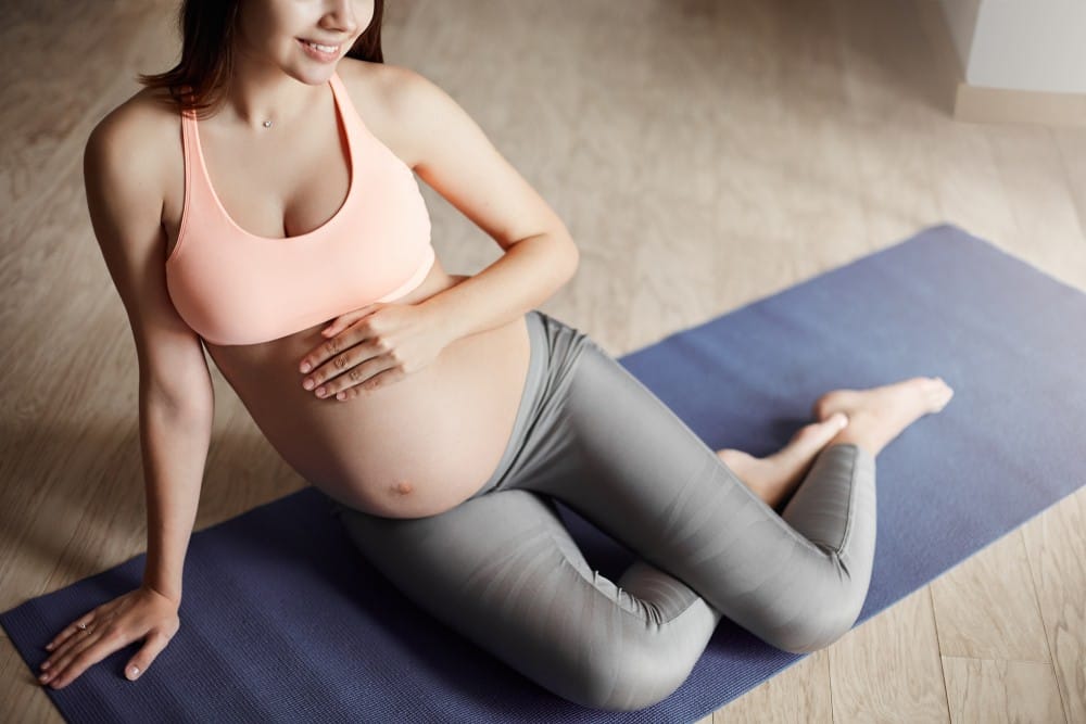 A woman in workout attire sitting on a yoga mat holding her pregnant belly