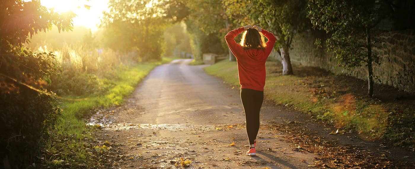 Woman in running gear walking with hands on head through forest path
