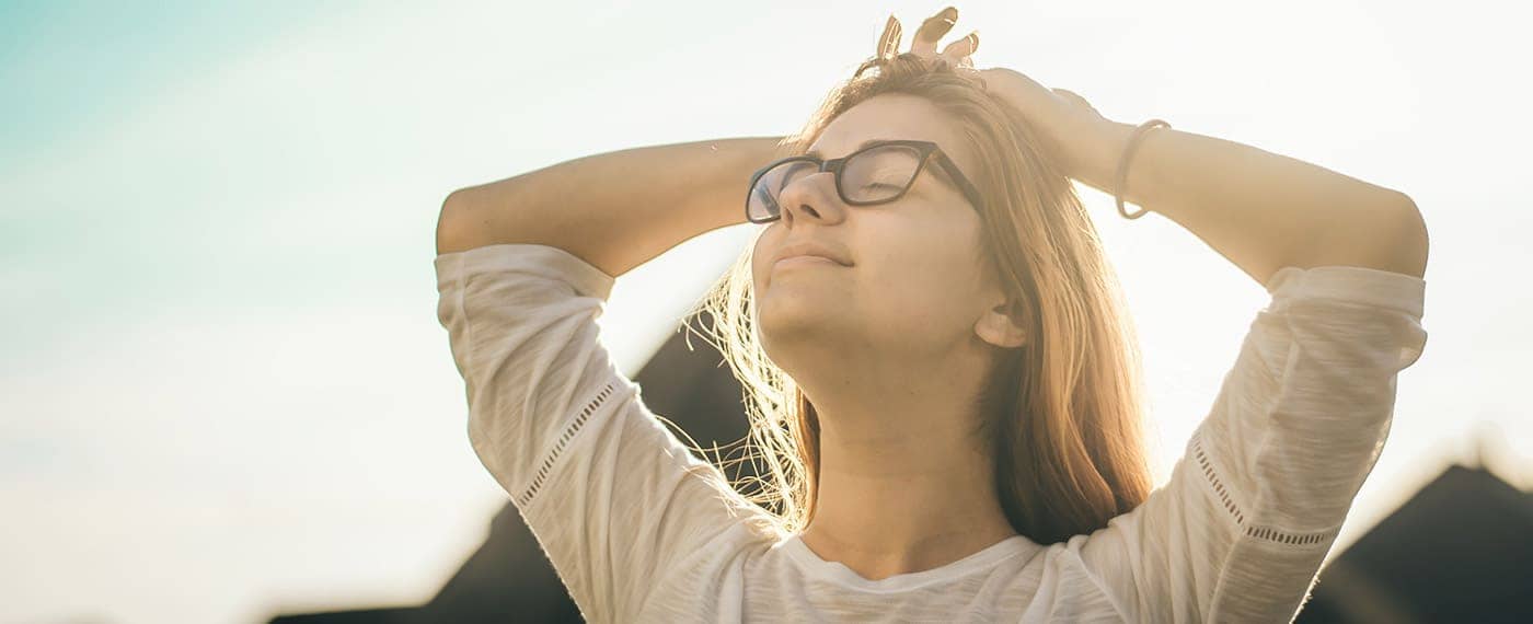 woman closing eyes and taking deep breathe with sunlight in the background to relieve stress