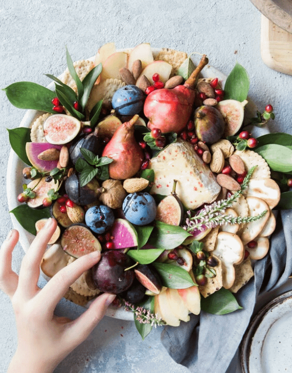 A large round bowl filled with fruit, crackers, nuts, and greens
